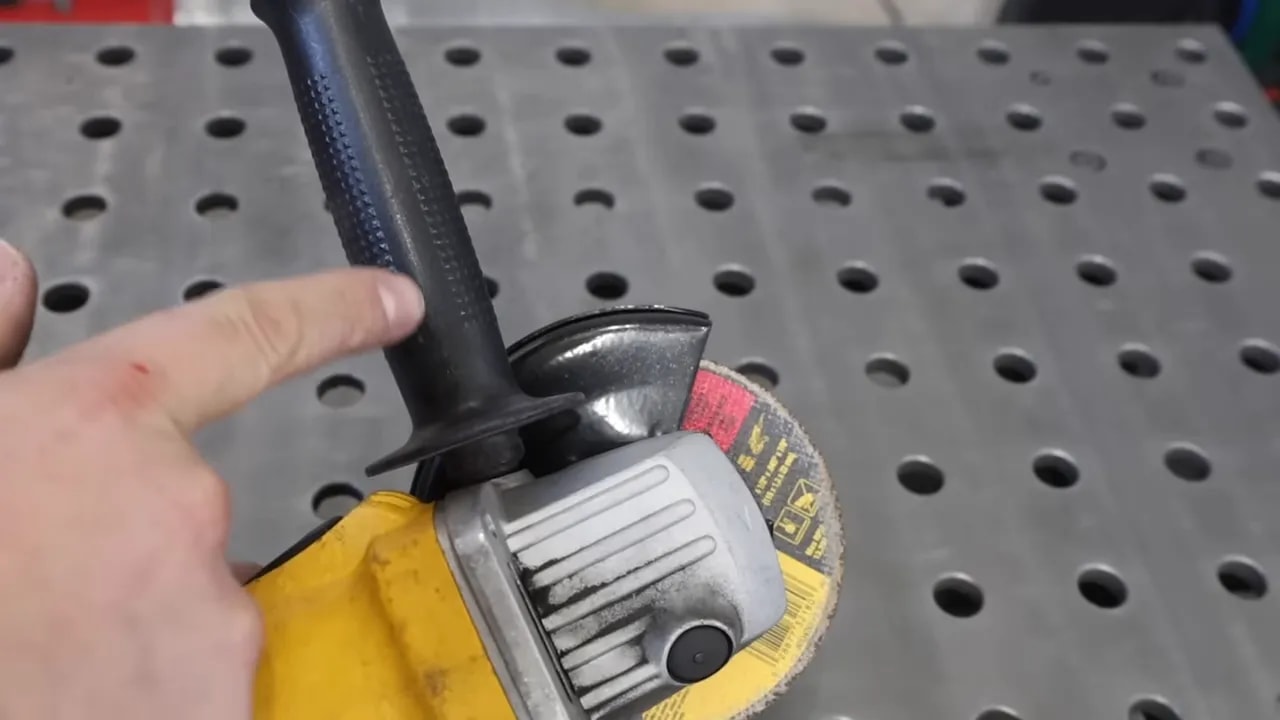 Finger pointing at the side handle of an angle grinder showing correct hand placement and the guard over the disc.